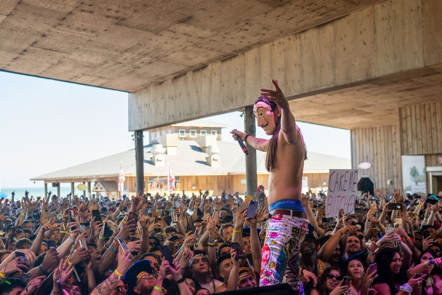 A shirtless, masked performer stands in front of a large audience at a beach.