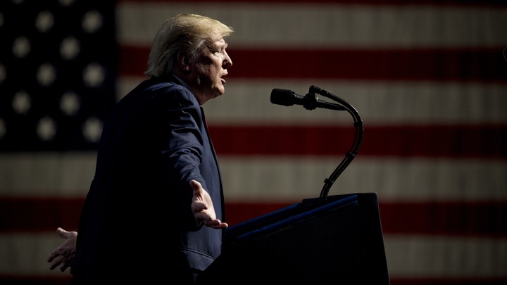 Donald Trump speaks into a microphone. An American flag hangs behind him.