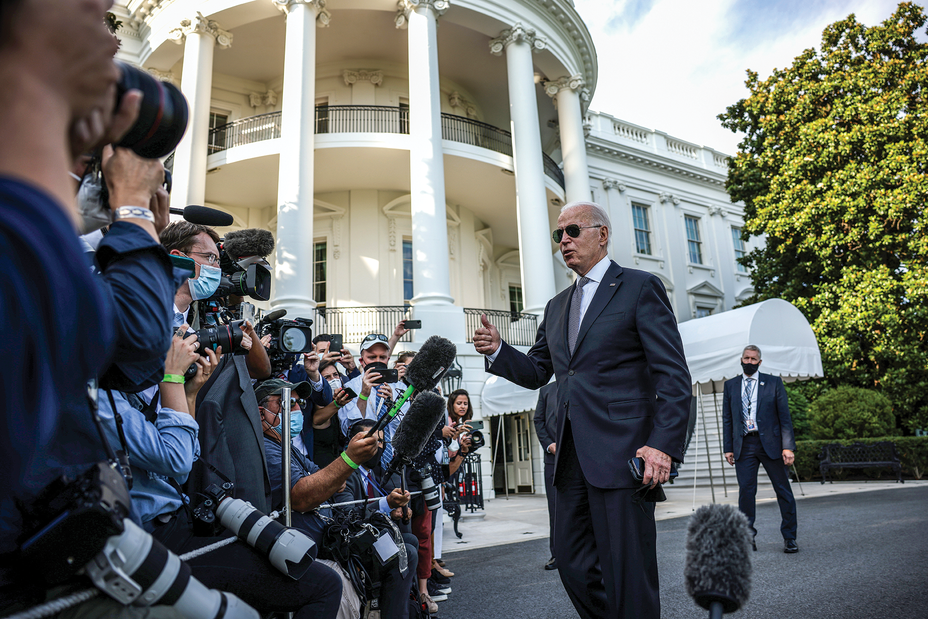 photo of Joe Biden in suit, tie, and aviator sunglasses talking with dense crowd of people with microphones and cameras outside columned White House portico