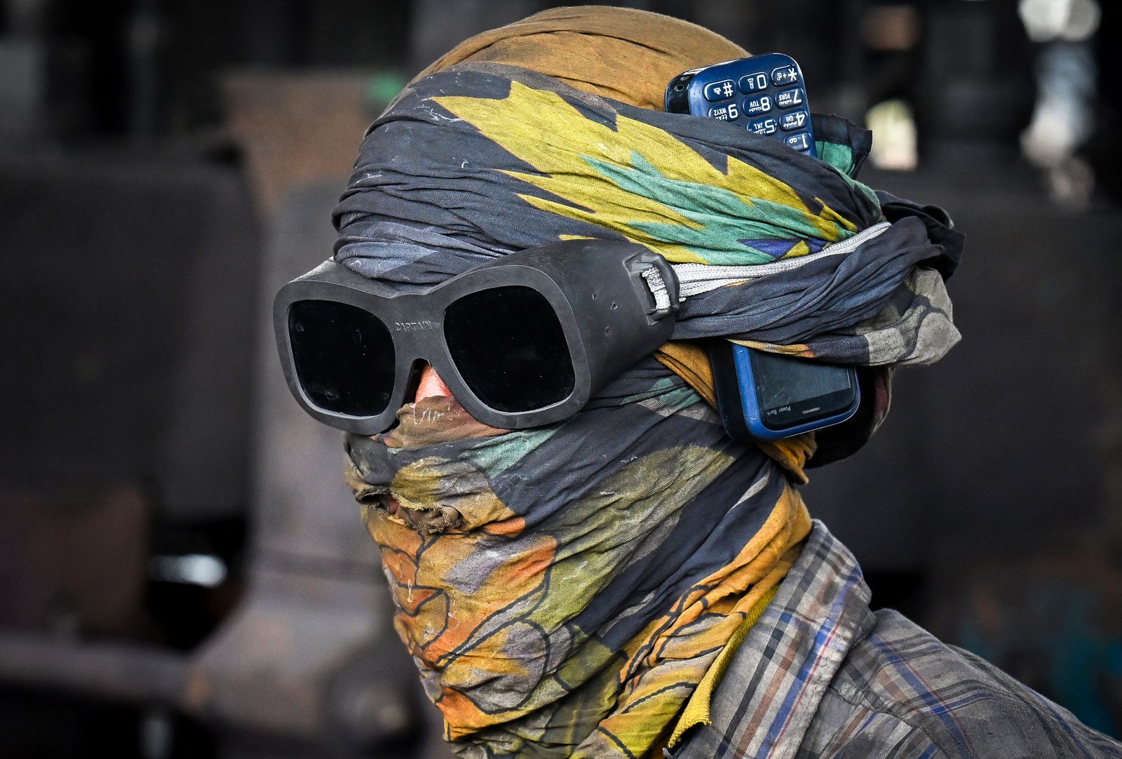 A worker wears a cloth wrapped around their head, as well as safety goggles, with a cell phone tucked into the folds of the cloth.