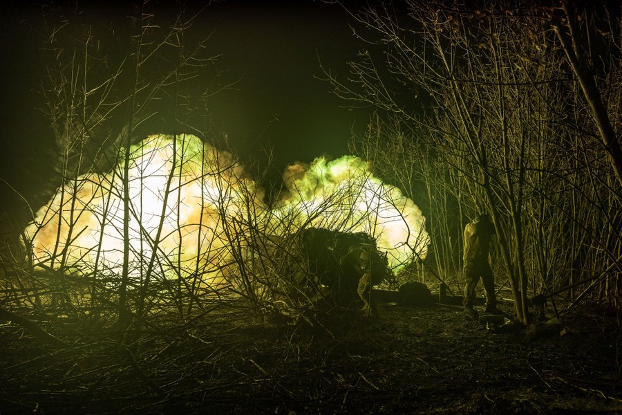 Soldiers fire a large piece of artillery at night, lit by a large muzzle flash.