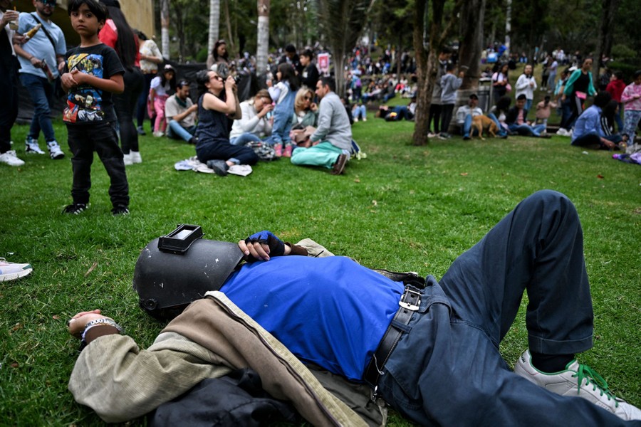 A person lies on their back on grass, wearing a welder's mask among a crowd of skygazers.