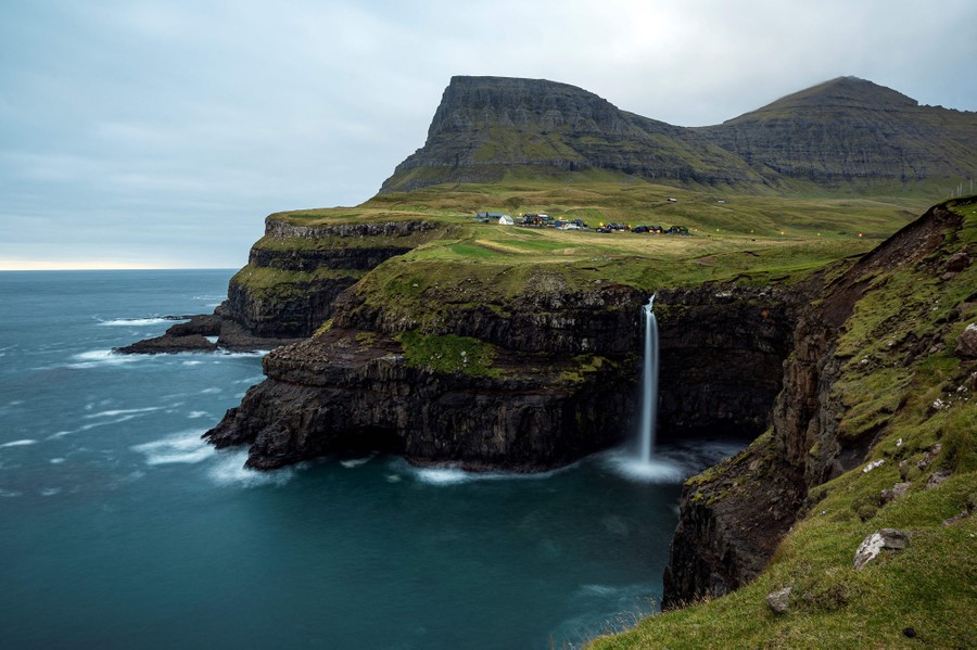 A rugged coastline of cliffs rises to rolling grass-covered hills, with a single dramatic waterfall plunging into the sea.