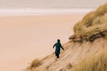 Photo of a person in a coat and hat scrambling along steep, grassy dunes next to a broad, sandy beach