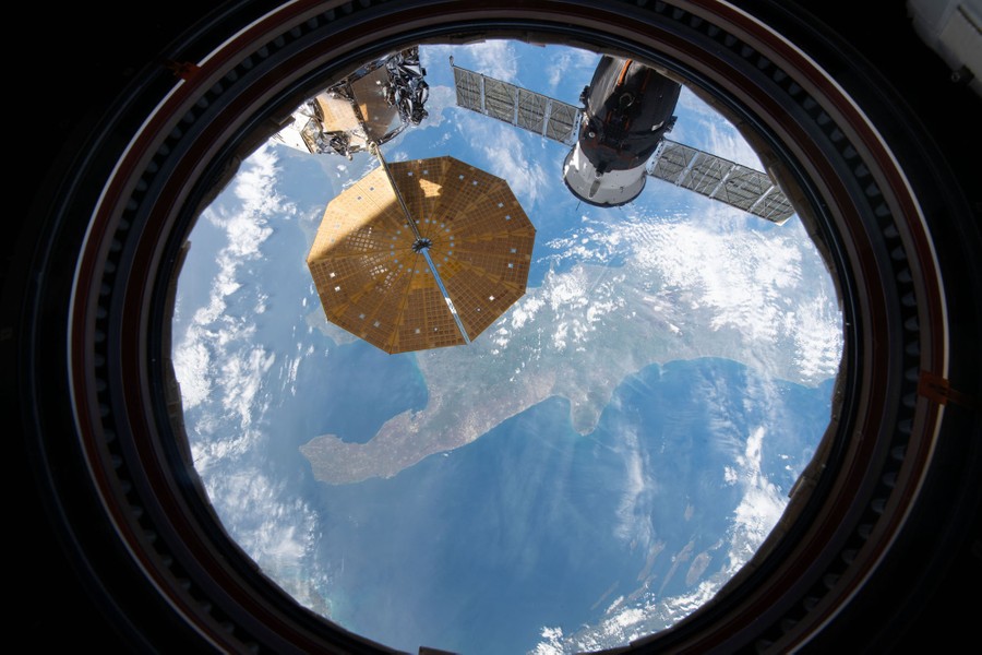 Parts of the ISS are seen in the foreground, with southern Italy far below, viewed through a window.