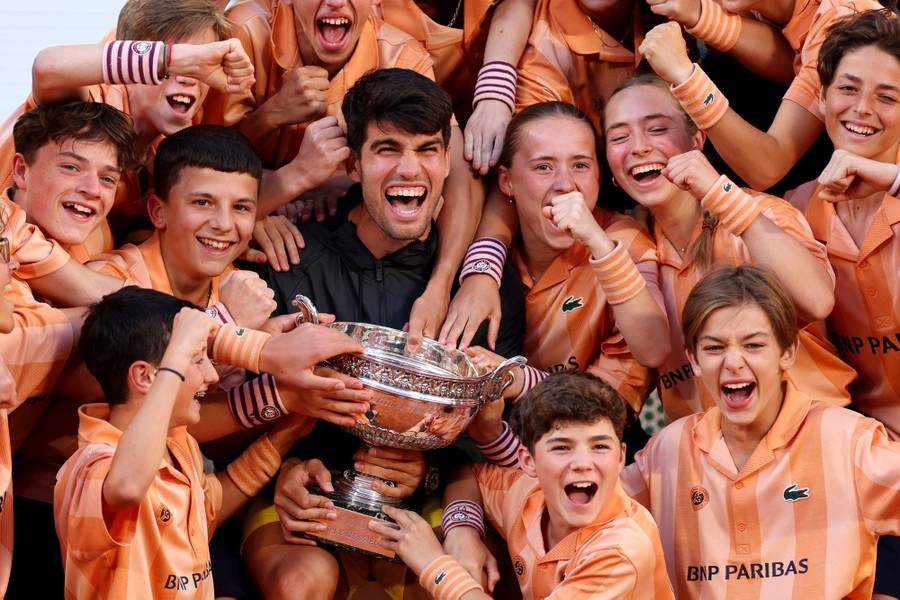 A tennis player smiles while sitting with a trophy among a crowd of excited young people.