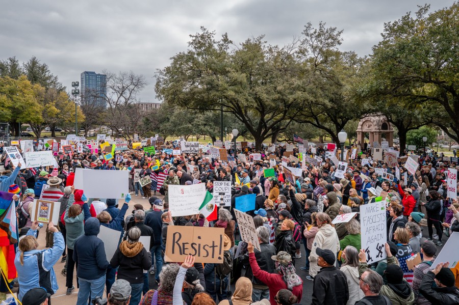 A crowd of people gather in a public space, holding protest signs, including one that reads 