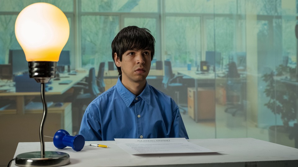 Julio Torres in a blue button down sitting at a table with a giant light bulb lamp
