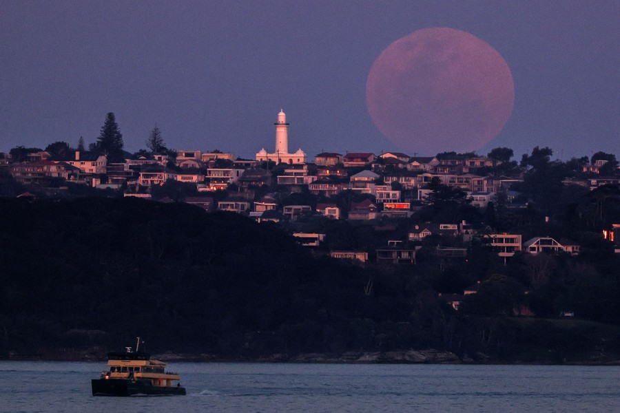 The full moon rises above a lighthouse and other houses, with a ferry in the foreground.