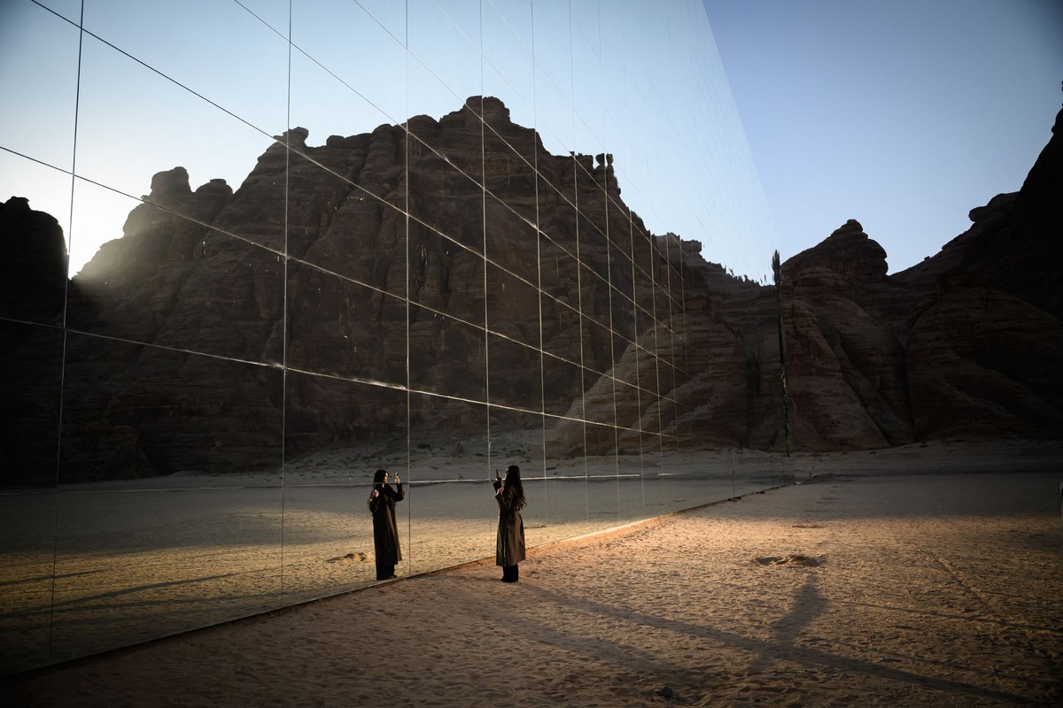 A person stands in a desert, facing the mirrored wall of a large building.