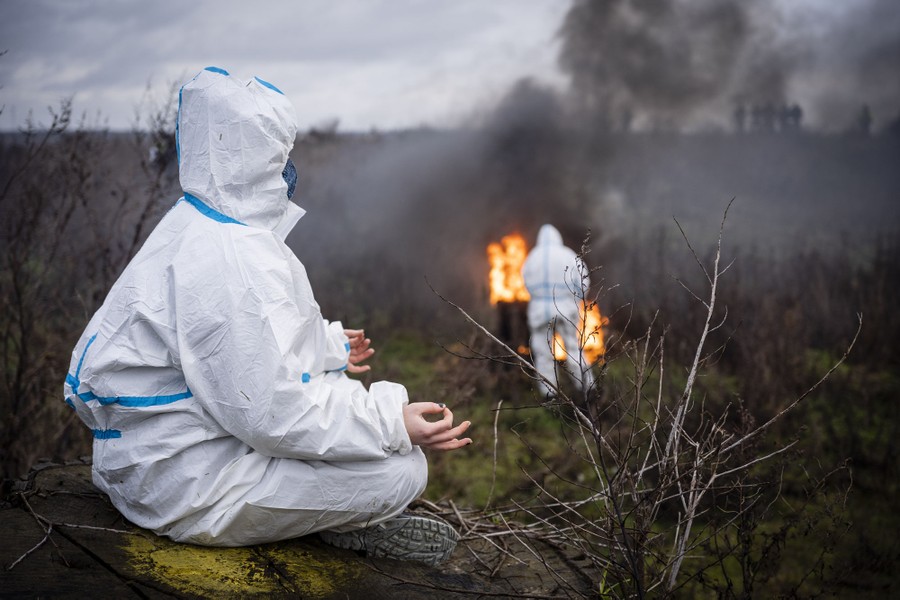 A person wearing white protective gear sits cross-legged, meditating, while another stands near a fire in the background.
