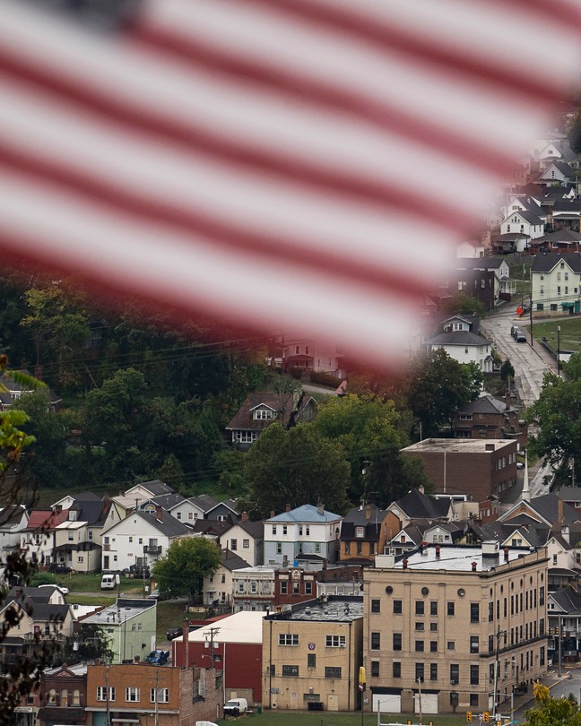 Color photograph of the town of Charleroi seen at a distance on what appears to be an overcast day. A blurry American flag whips across the top of the frame at close range.