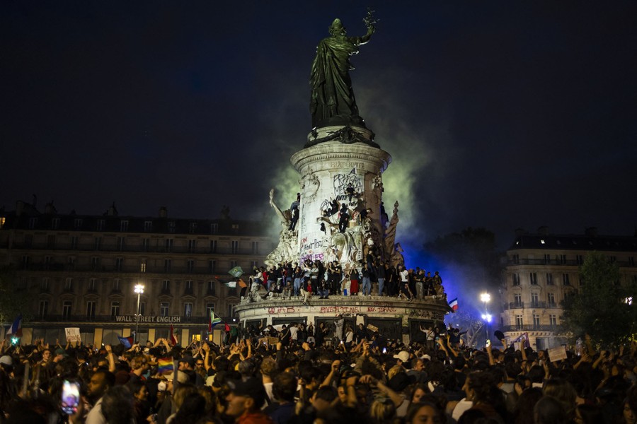A crowd of people demonstrates around and on a tall statue in a city square.