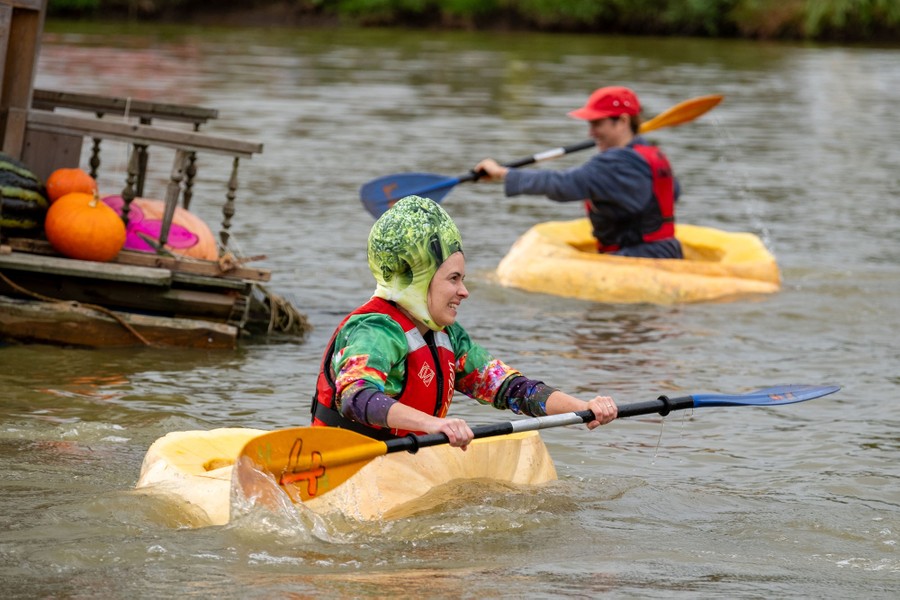 Two people paddle in a lake, floating in huge hollowed-out pumpkins.
