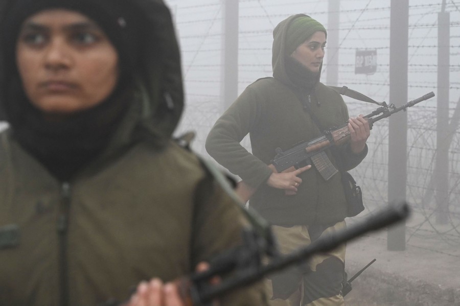 Two people carry rifles, walking along a border fence in the fog.