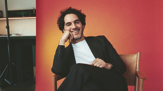 A color photograph of Benny Safdie seated in front of a red backdrop