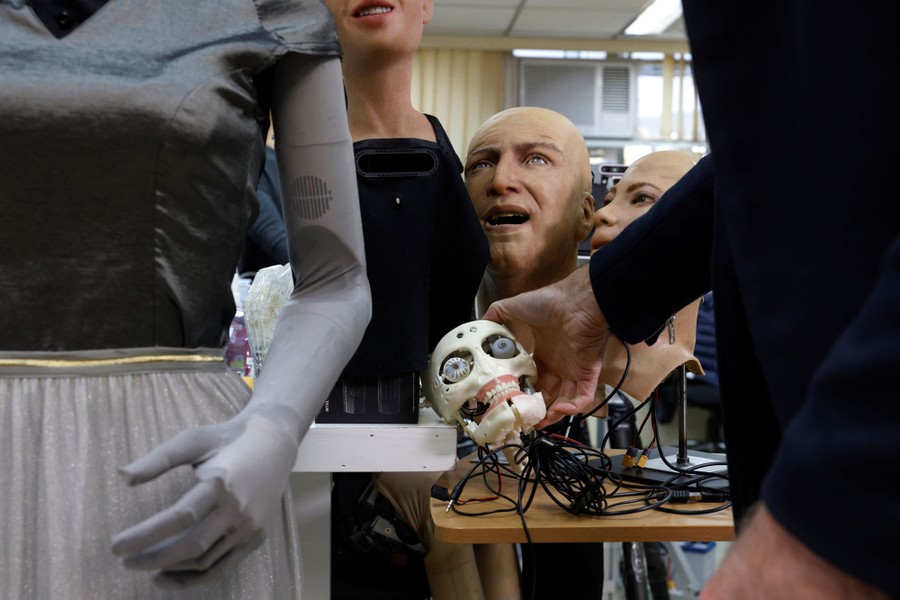 Several lifelike robotic human heads are seen around a hand holding a small plastic, robotic skull.