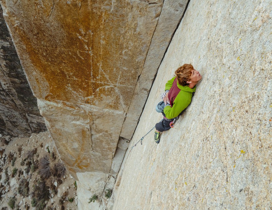 A rock climber rests on a narrow ledge while climbing a very flat cliff.