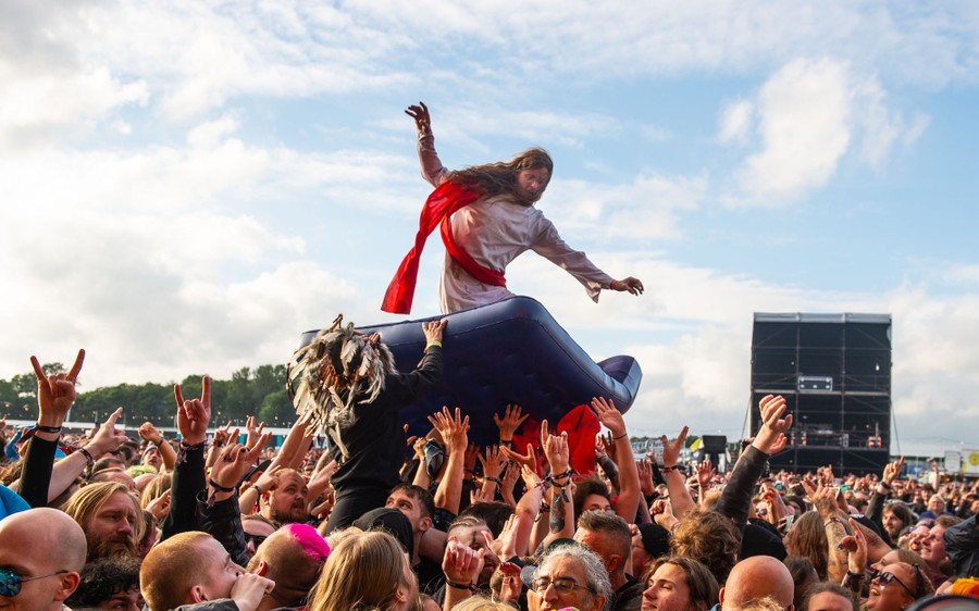 A person dressed as Jesus stands atop a mattress that is held aloft by dozens of fans in a large crowd.