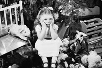 A girl sits with her face in her hands, looking disgruntled, surrounded by gifts under a Christmas tree