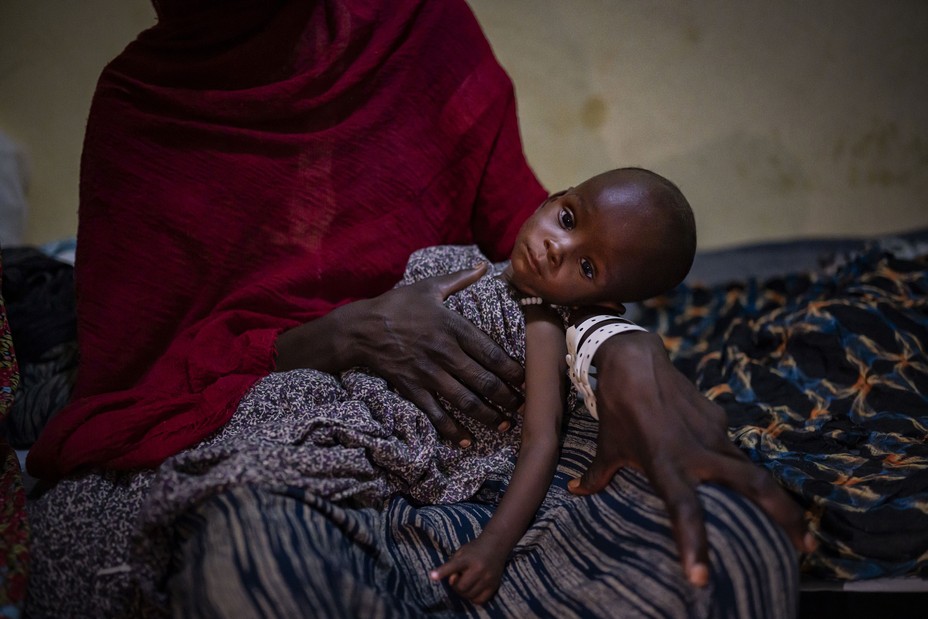 photo of woman from shoulders down wrapped in red shawl and holding emaciated baby in her lap, sitting on brightly patterned blanket