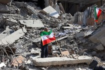 Photograph of a man holding up an Iranian flag atop the wreckage of a bombed building