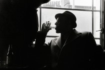 A man wearing a hat and a wool coat drinks from a glass while he sits next to a window