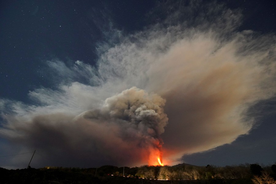 A distant view of ash and lava being thrown skyward from an erupting volcano.