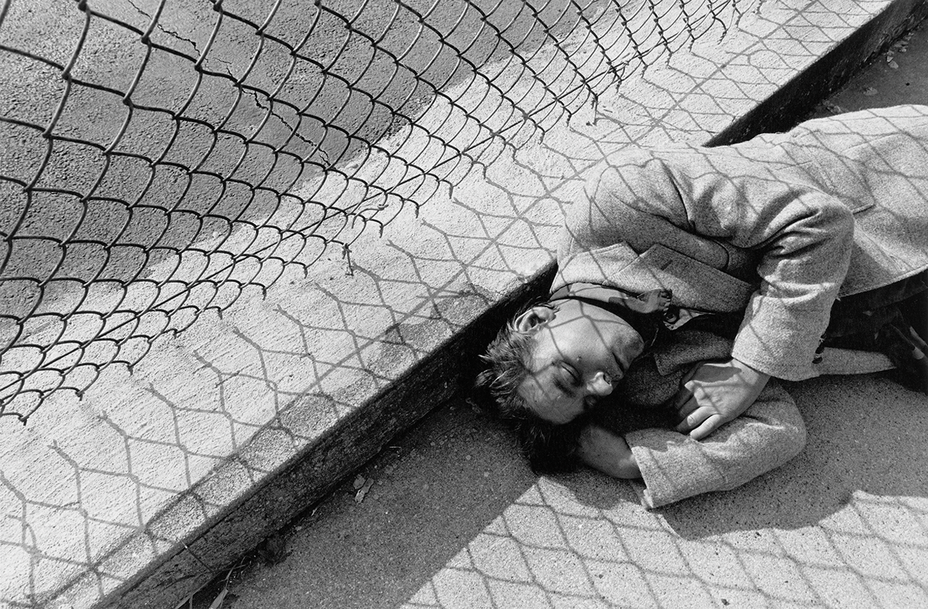 black-and-white photo of man lying with eyes closed next to chain-link fence that casts patterned shadow over him