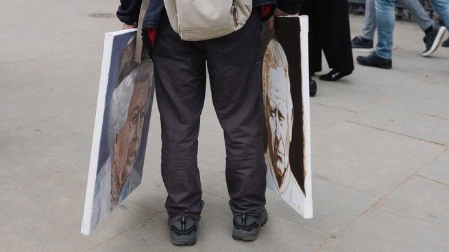 Someone shown from the legs down holds two paintings, one of Queen Elizabeth II and one of King Charles III