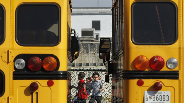 Preschool students play behind a fence. They are framed between two yellow school buses.