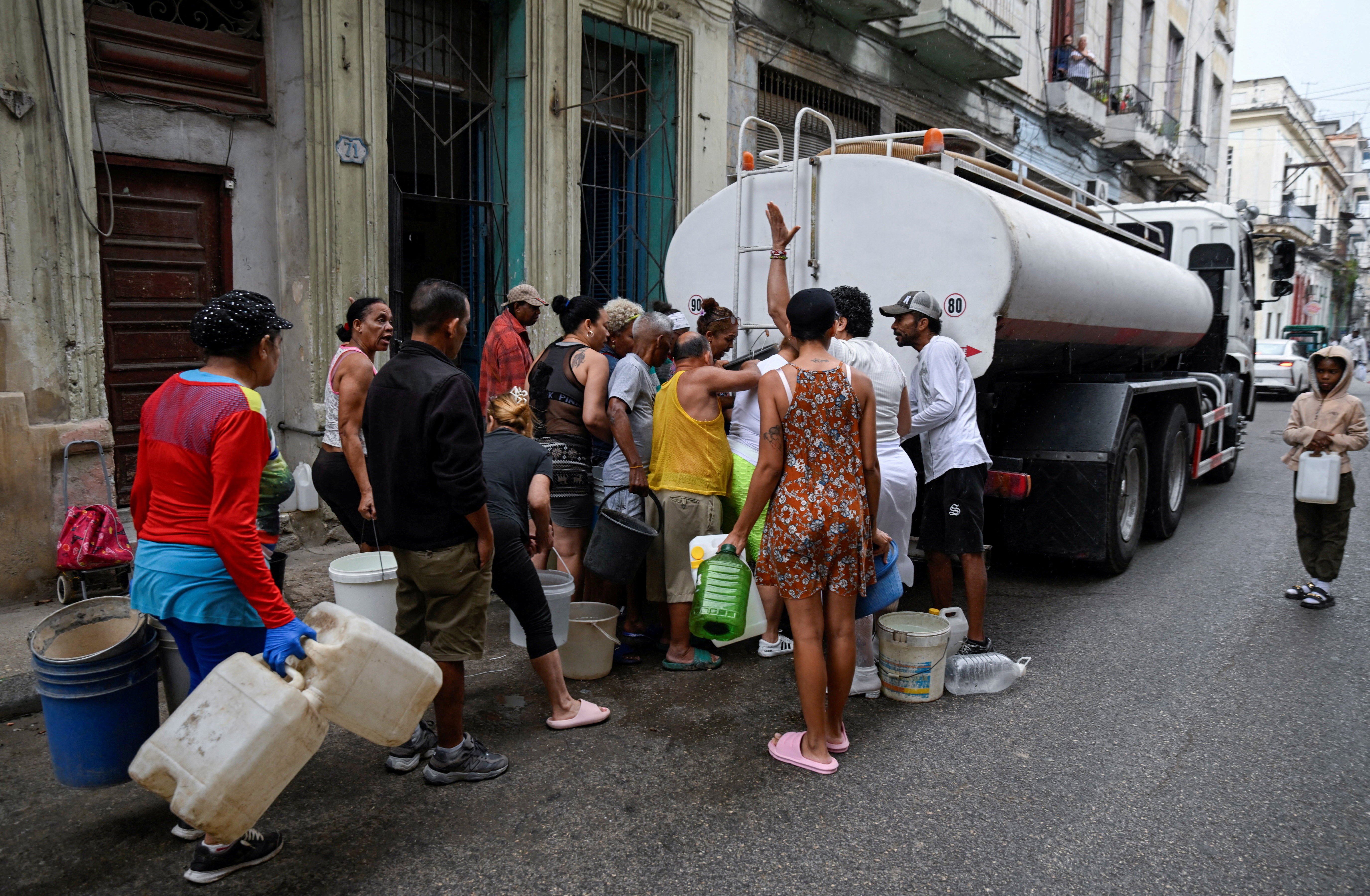 People gather around a water tanker truck to fill up buckets and other containers in a street.