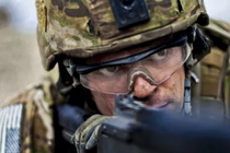A person in camouflage clothing and helmet, wearing safety goggles, peers down the barrel of a gun.