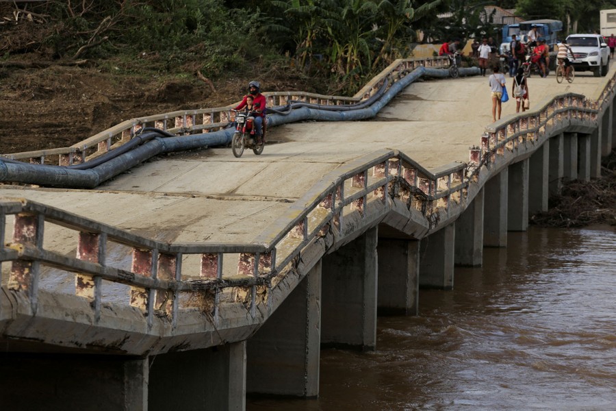 People walk and drive across an uneven flood-damaged road bridge.