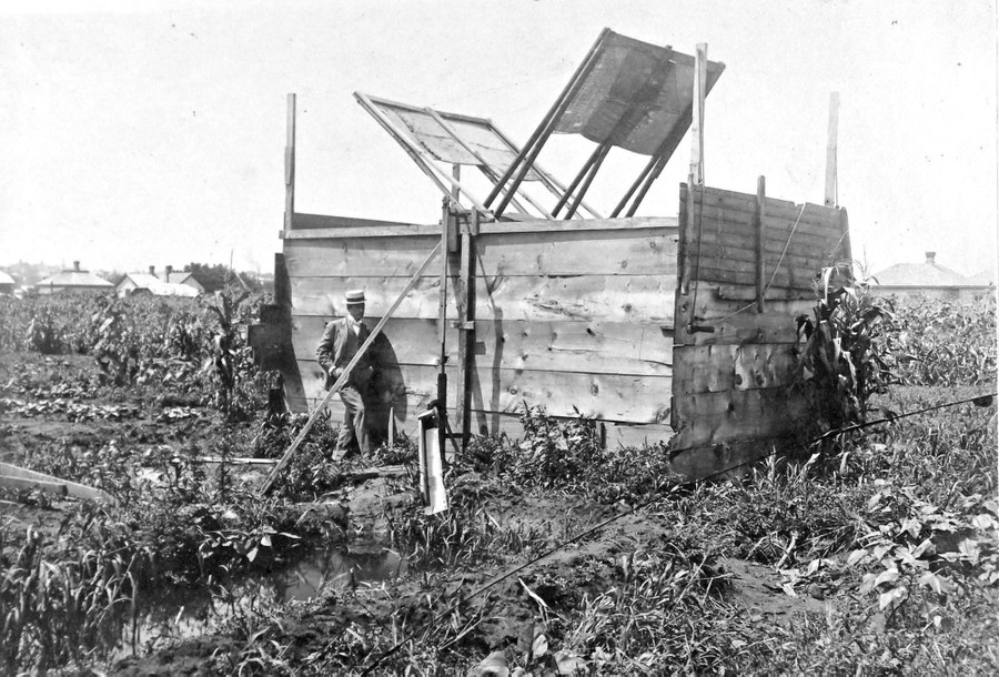 A man stands beside a large wooden structure in a farm field, with a sort of paddle-wheel style of a windmill inside the structure.