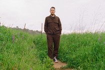 Color photo of man in brown shirt and pants standing with hands in pockets on trail surrounded by green grass and wildflowers.