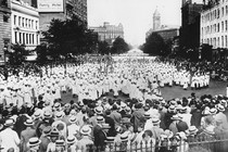 A large crowd of people wearing white gowns and white, pointed hats approaches a fence, against which stand other people in regular clothes.