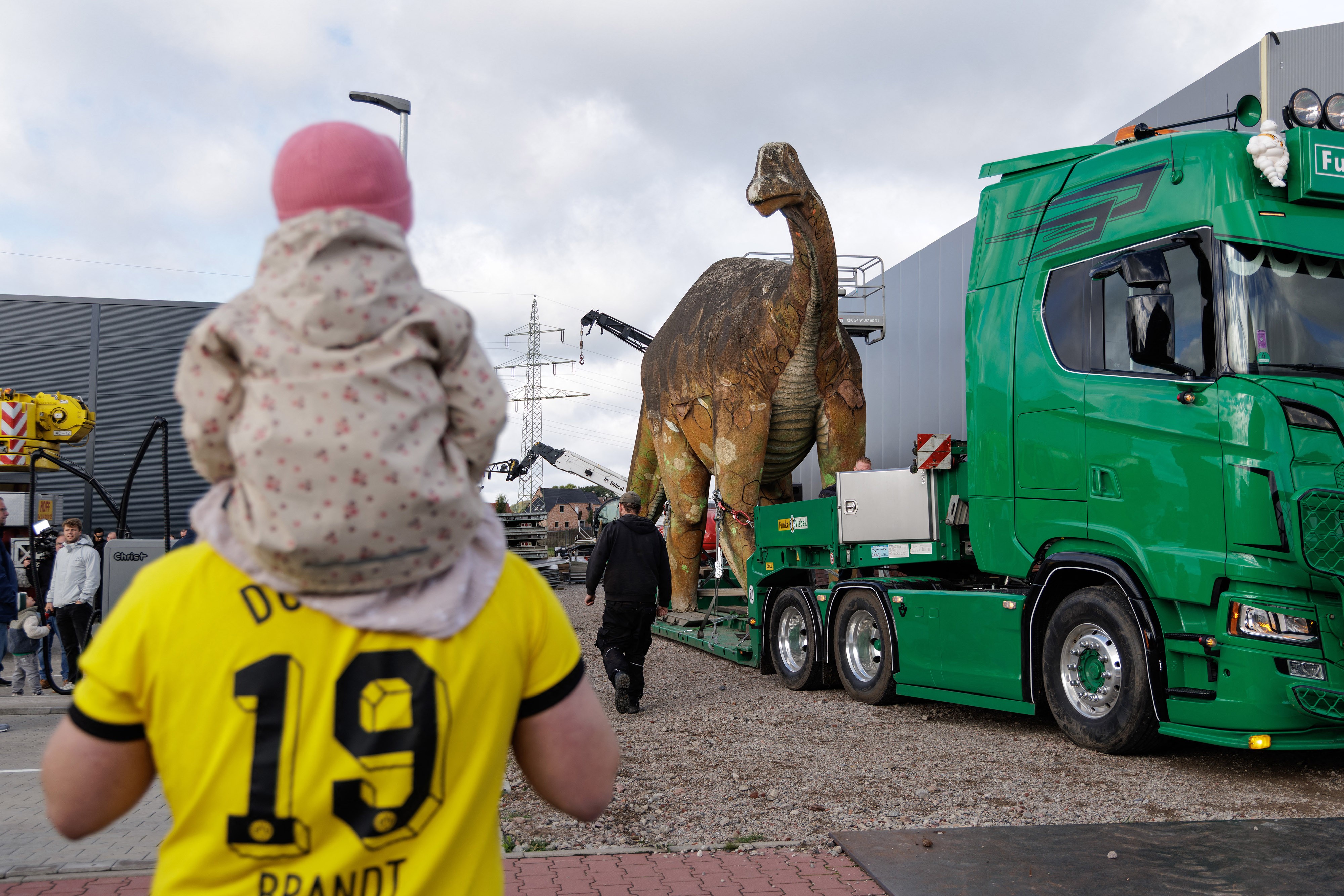 A life-sized dinosaur statue is loaded onto the low bed of a truck, as people watch.