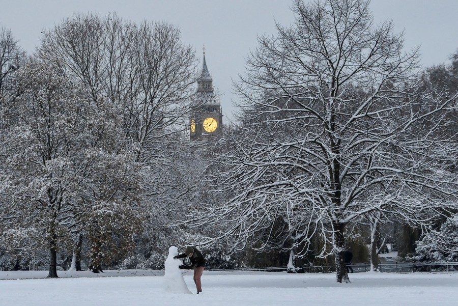 A person builds a snowman in a park in London with snow-covered trees and Big Ben in the background.