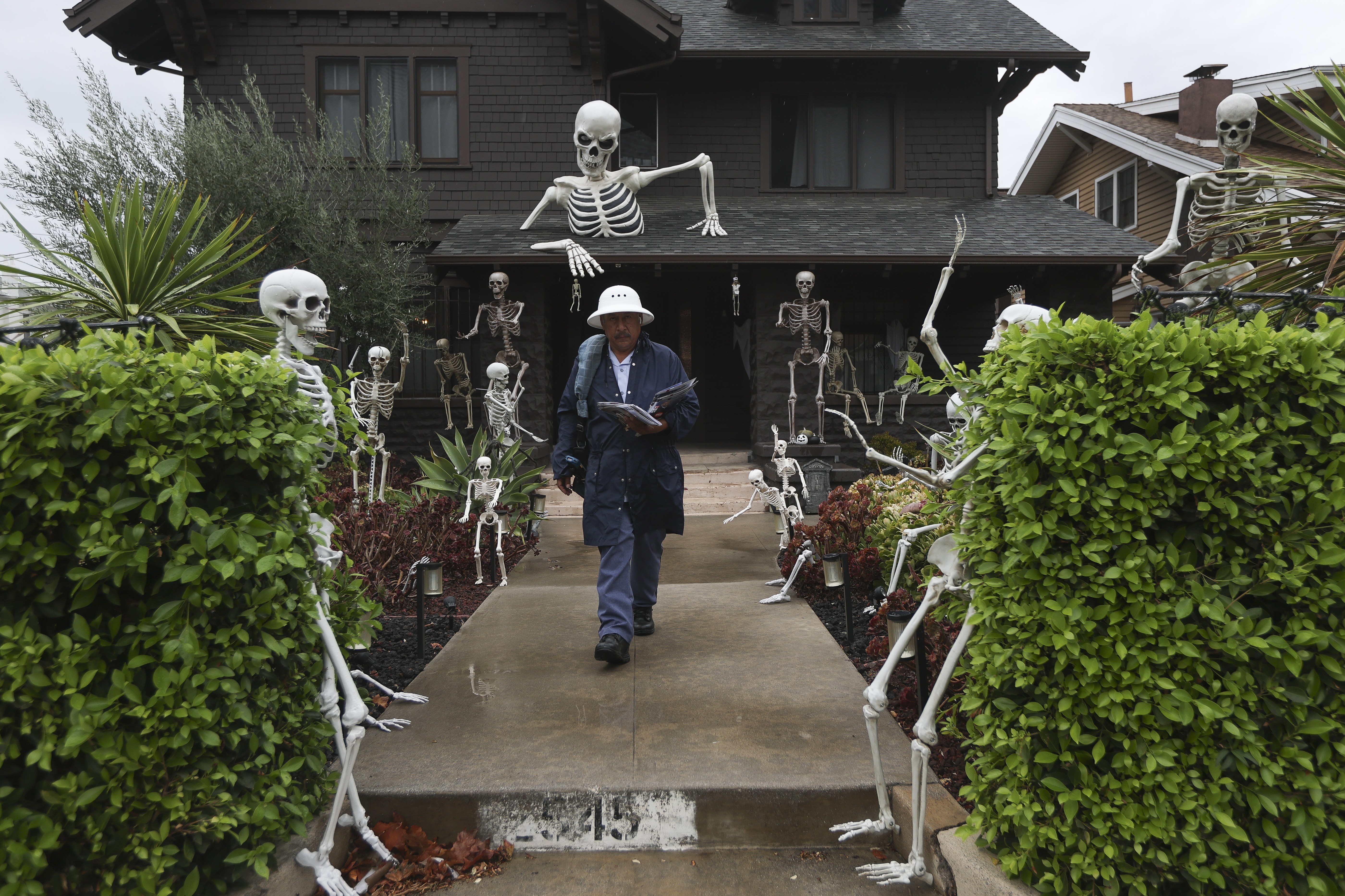 A mail carrier walks down a path of a house decorated with many Halloween skeleton displays.