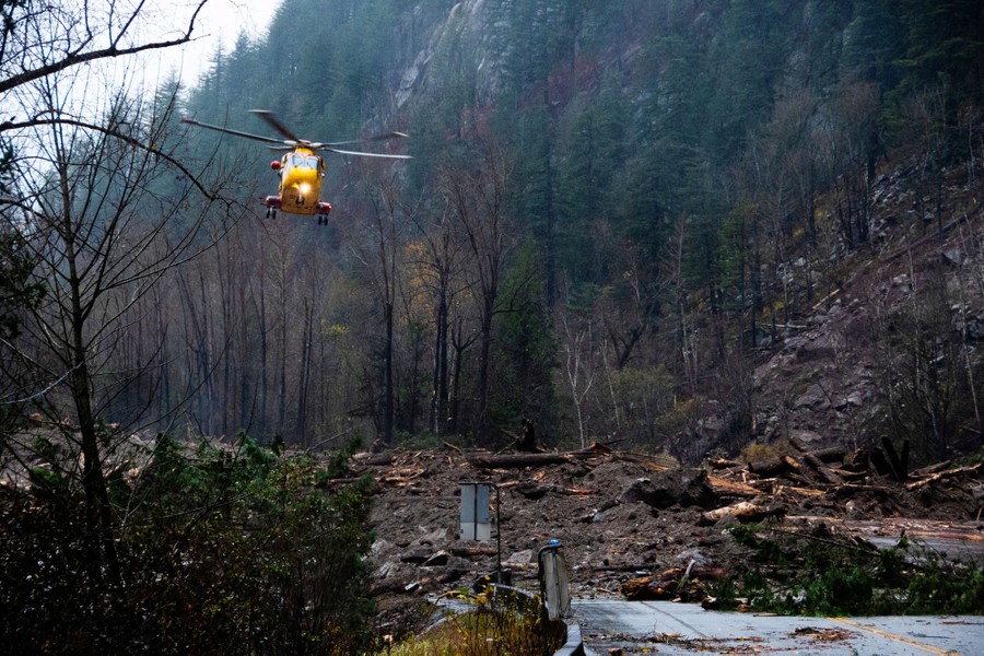 A helicopter descends into a narrow mountain valley, above a mudslide-covered road.