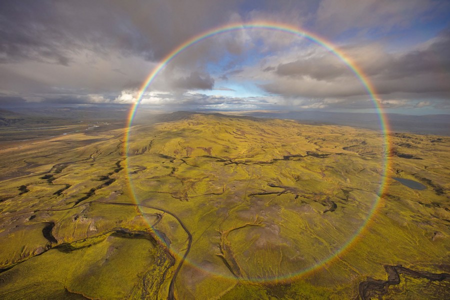 An aerial view of treeless hills beneath a partly cloudy sky, seen with a rainbow forming a full circle