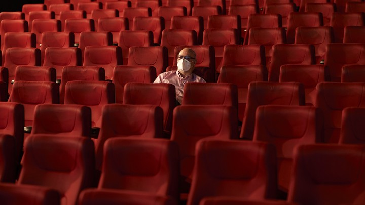 A man wearing a mask sits alone in a theater.