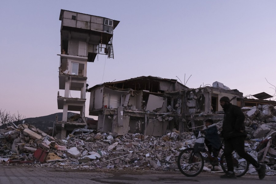 A man walks beside his son on a bicycle as they pass a heavily damaged building, including one tall section that stands precariously.