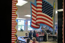 The photograph peers into an elementary school classroom through a door with an American flag sticker on it. 