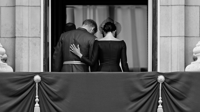 A black and white photograph of Harry and Meghan returning to the inside of Buckingham Palace from a viewing balcony.