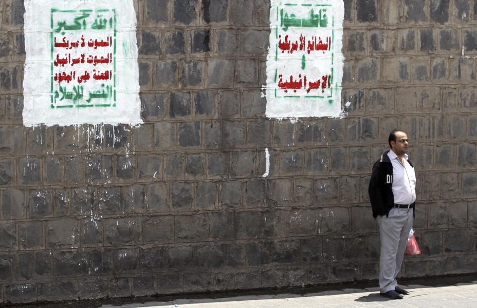 a man stands on a street with Houthi slogans painted on a wall