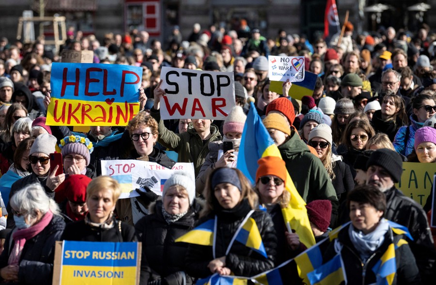 A crowd of protesters hold up handwritten signs, with two that read "Help Ukraine" and "Stop War."