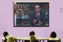 A projector screen is filled with a screengrab from John Oliver's show. The word "school" appears behind Oliver.