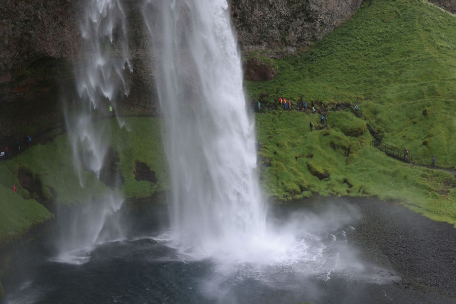 People walk on a path along a cliff beneath a large waterfall.